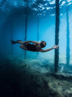 Serbest dalgıç kız, mavi okyanustaki iskelede yüzgeçleriyle süzülüyor. Dişi, iskele sütunları arasında su altında yüzüyor.