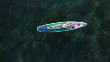 14 Nisan 2024. Menorca, İspanya. Sessiz denizde ayağa kalkmış kürek çeken kadın. Red paddle sup tahtasının üzerinde dinleniyor. Hava görünümü