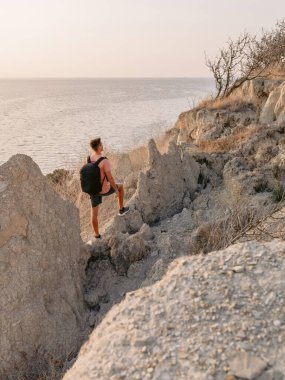 Sırt çantalı yakışıklı adam sıcak güneş ışığıyla kayalık okyanus kıyısında kalıyor..