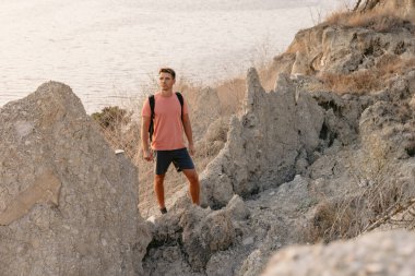 Sırt çantalı yakışıklı adam sıcak güneş ışığıyla kayalık okyanus kıyısında kalıyor..