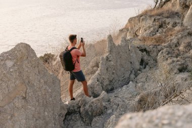 Deniz kıyısındaki kayalıklarda gün batımını seyreden yürüyüşçü. Açık havada akıllı telefonu olan bir gezgin..
