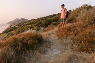 Sırt çantalı gezgin sıcak güneş ışığıyla okyanus kıyısında yürüyor..