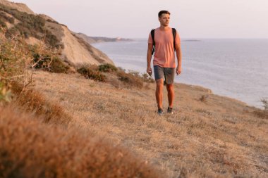 Sırt çantalı bir yürüyüşçü sıcak güneş ışığıyla kıyı şeridinde yürüyor..
