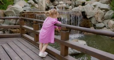 Yaz parkında şık pembe elbiseli küçük bir kız çocuğu ve şelaleye bakıyor. Yüksek kalite 4k görüntü