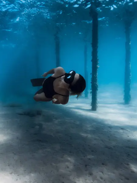 stock image Free diver girl glides with fins dive under the pier in blue ocean. Female swims underwater dives near the pier pillars