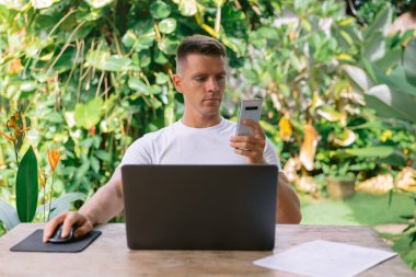 Ciddi bir adam akıllı telefon tutuyor ve açık bir planlama ofisinde dizüstü bilgisayarla çalışıyor..