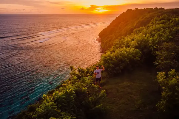 Bali 'de gün batımında okyanus kıyısında elini sallayan bir adam. Drone görünümü