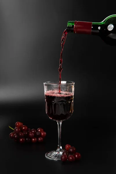Red wine is poured into a wine glass on a black background with grapes and splash copy space