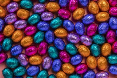 Pile of shiny wrapped chocolate Easter eggs, in colors red, blue and golden,green and purple. Festive Easter concept, top view pink background Happy easter close-up candy sweets concept stack