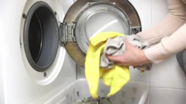 Close Up of a Beautiful Young Woman in Homely Clothes Sitting in Front of a Washing Machine. She Loads the Washer with Dirty Laundry. Bright and Spacious Living Room with Modern Interior. 4k