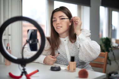 Güzellik blogları, teknoloji ve insanların konsept portresi mutlu gülümseyen bir kız blogcunun telefon ışığı ve akıllı telefonuyla evde makyaj yapması. fırça kullanarak etkili bir video hazırlamak