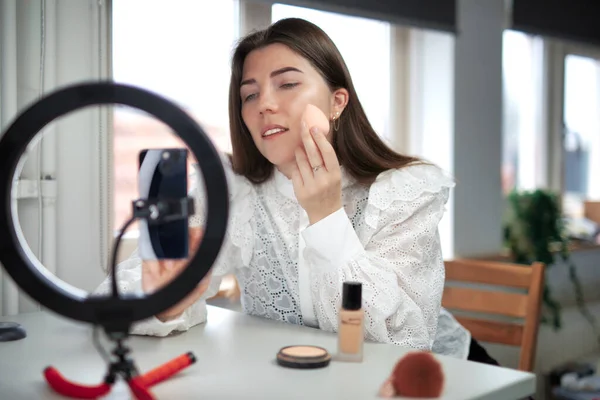 beauty blogging, technology and people concept portrait of a happy smiling girl blogger with ring light and smartphone applying make up at home. making a influencer video Using brush