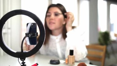 Güzellik blogları, teknoloji ve insanların konsept portresi mutlu gülümseyen bir kız blogcunun telefon ışığı ve akıllı telefonuyla evde makyaj yapması. fırça kullanarak etkili bir video hazırlamak