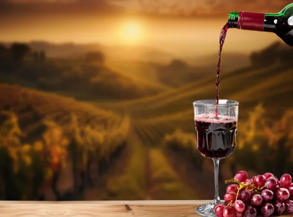 Red wine on wooden table and vineyard in green Tuscany sunset, Italy blurred background, wine glass and grapes beautiful landscape of grapes field copy space