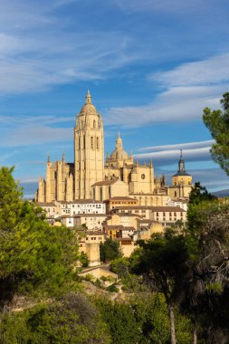 Segovia Katedrali 'nin yeşil ağaçlar arasındaki görüntüsü, ortaçağ mimarisi ile çevrili, gökyüzünde merceksi bulutlar.