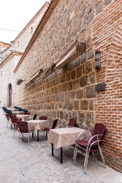 Medieval narrow cobbled street in Avila, Spain, with restaurant tables and dining area, no people.