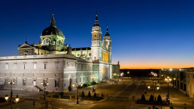 Madrid, İspanya, 09.10.21. Almudena Katedrali (Santa Maria la Real de La Almudena) ve Plaza de la Armeria gece görüşü, günbatımından sonra aydınlanan binalar, mavi ve turuncu gökyüzü.