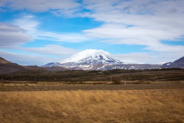 Icelandic landscape with grasslands, lava fields and snowcapped Hekla volcano with peak covered in clouds, blue sky. clipart