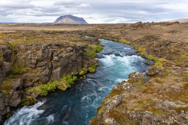 Thjorsardalur valley, South Iceland, landscape with turquoise glacial wild river, volcanic rock formations overgrown with moss and grass and mountains in the background. clipart