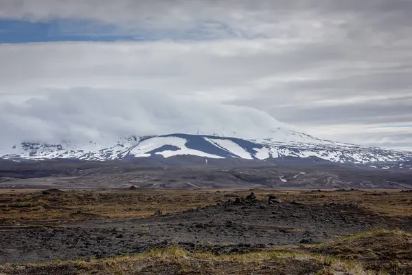 Raw Icelandic landscape with volcanic lava field and snowcapped Hekla volcano with peak covered in clouds.