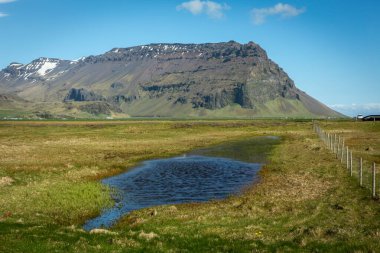 Güneşli bir günde, arka planda yeşil çayırlar, bataklıklar ve kayalık dağlar olan Güney İzlanda Eyjafjoll sıradağları manzarası.