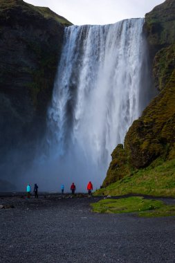 Skogafoss, İzlanda, 16.05.2022. Su püskürten Skogafoss şelalesi manzarası ve renkli ceketli insanlar aşağıda yürüyor..