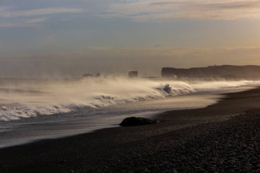 Reynisfjara Kara Kum Sahili 'ne çarpan spor ayakkabı dalgaları. Güçlü rüzgarlar esiyor. Su ve volkanik siyah kum, gün batımı, aşırı hava, İzlanda..