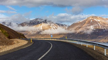 Asfalt yol 94 İzlanda kıyısı boyunca uzanan çimenli dağlarla kaplı arka planda bulutlar ve karla kaplı.