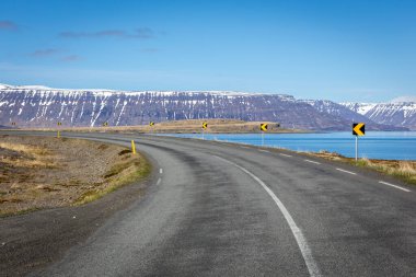 İzlanda 'daki Westfjords' a giden asfalt yolda kar kaplı dağlar ve arka planda deniz manzarası var..