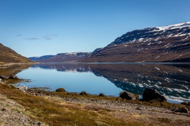 Batı fiyortları İzlanda kıyı şeridi. Karla kaplı dağlar kristal berrak sulara, mavi gökyüzüne yansıyor..