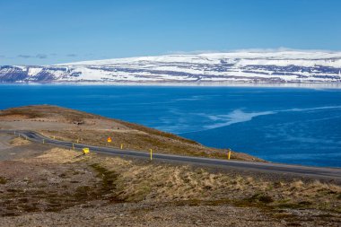 İzlanda 'daki Westfjords' a giden asfalt yolda kar kaplı dağlar ve arka planda deniz manzarası var..