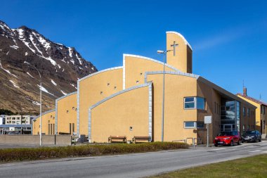 Isafjordur, Iceland, 23.05.22. Isafjardarkirkja church in Isafjordur, Westfjords, Iceland. Modern architecture church with yellow facade and snowcapped mountains in the background. clipart