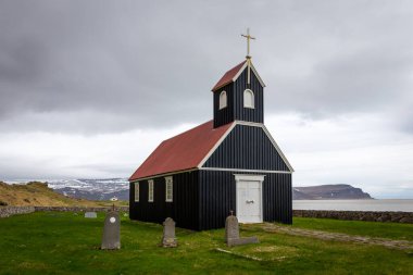 Sauraejar, İzlanda, 24.05.22. Sauraejarkirkja (Sauraejar Kilisesi) siyah ahşap cephe, kırmızı çatı ve mezarlık, arka planda Raudisandur sahili, Westfjords, İzlanda.