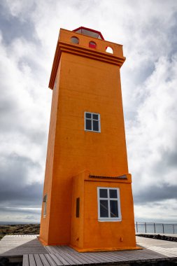 Svortuloft Lighthouse (Svortuloftsviti), bright orange lighthouse on the westernmost tip of Snaefellsnes peninsula, Iceland with wooden walking platforms for tourists and black volcanic rocks around. clipart