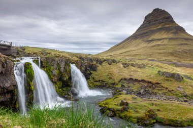 Mt. Kirkjufell and Kirkjufellsfoss waterfall landscape, spring, long exposure view with mountain covered with moss on a cloudy day, Snaefellsnes Peninsula, Iceland. clipart