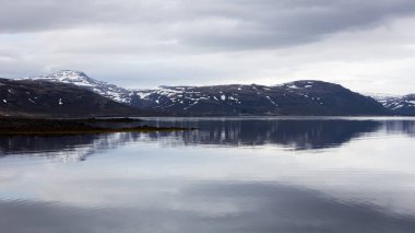 İzlanda 'da Flokalundur çevresindeki Westfijords kıyı şeridi sakin deniz yüzeyi ve karla kaplı dağlarla suya yansıyan bulutlu bir gün..