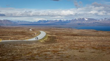 Winding tarmac road 36 with cars driving along Thingvallavatn lake in Thingvellir National Park, Iceland, lava fields and snowcapped mountain in the background. clipart