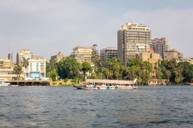 Giza, Mısır, 25.07.21. Tahta tur teknesi Alhaga 'da Nil nehri boyunca arka planda Giza şehri manzarası olan insanlar, bakımsız konut binaları ve yemyeşil bitki örtüsü ve tekne rıhtımı..