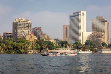 Giza, Mısır, 25.07.21. İhmal edilmiş konut binaları, Sheraton Oteli, Nil Nehri 'nde insanlarla dolu bir tur botu ve yemyeşil bitki örtüsü olan Giza şehri kıyısı silueti..