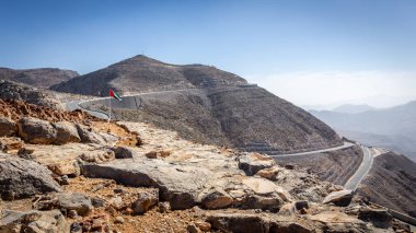 Jebel Jais dağ zirvesi kıvrımlı asfalt yolu yamacı kesiyor ve rüzgarda uçan BAE ulusal bayrağı, Ras al Khaimah, Birleşik Arap Emirlikleri.