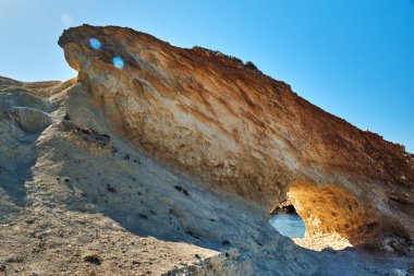 Kayanın üzerine doğal bir kemer oyulmuş engebeli bir kaya oluşumu. Açık mavi gökyüzünün altında parlak güneş ışığıyla aydınlatılıyor..
