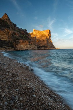 Mavi denizin üzerinde yükselen kaya, batan güneşin turuncu ışınlarıyla mavi gökyüzünün arka planına doğru yükselir.