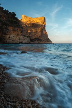 Mavi denizin üzerinde yükselen kaya, batan güneşin turuncu ışınlarıyla mavi gökyüzünün arka planına doğru yükselir.