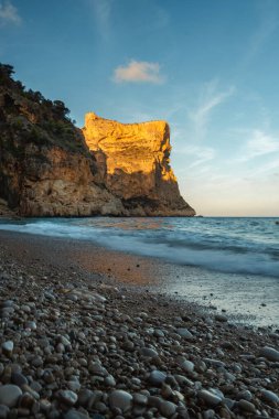 Mavi denizin üzerinde yükselen kaya, batan güneşin turuncu ışınlarıyla mavi gökyüzünün arka planına doğru yükselir.