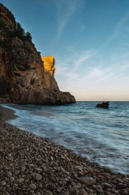 Mavi denizin üzerinde yükselen kaya, batan güneşin turuncu ışınlarıyla mavi gökyüzünün arka planına doğru yükselir.