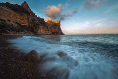 Mavi denizin üzerinde yükselen kaya, batan güneşin turuncu ışınlarıyla mavi gökyüzünün arka planına doğru yükselir.