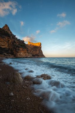 Mavi denizin üzerinde yükselen kaya, batan güneşin turuncu ışınlarıyla mavi gökyüzünün arka planına doğru yükselir.