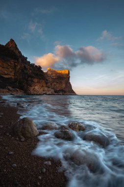 Mavi denizin üzerinde yükselen kaya, batan güneşin turuncu ışınlarıyla mavi gökyüzünün arka planına doğru yükselir.