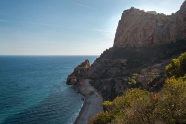 Mavi denizin üzerinde yükselen kaya, batan güneşin turuncu ışınlarıyla mavi gökyüzünün arka planına doğru yükselir.