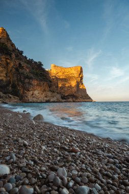 Mavi denizin üzerinde yükselen kaya, batan güneşin turuncu ışınlarıyla mavi gökyüzünün arka planına doğru yükselir.